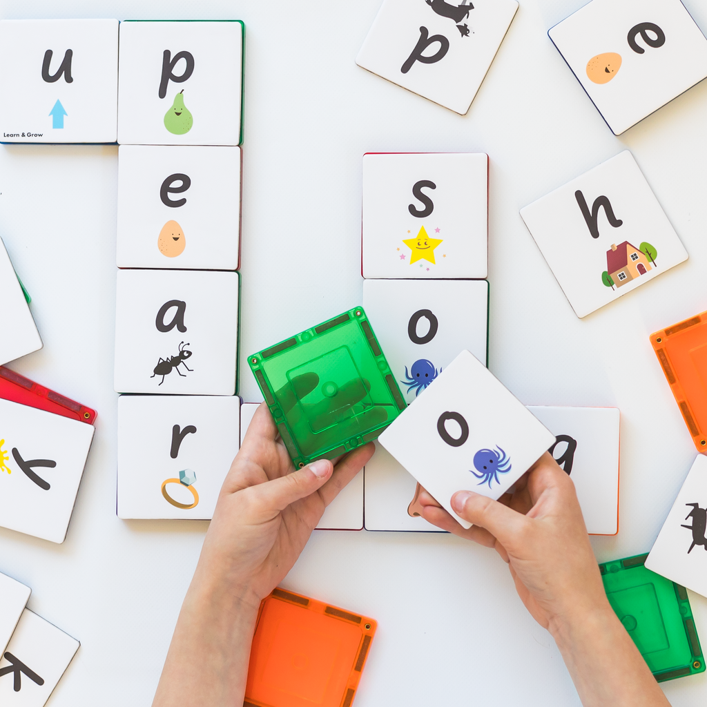 Child holding green magnetic tile with lowercase o topper next to it with toppers on table in background